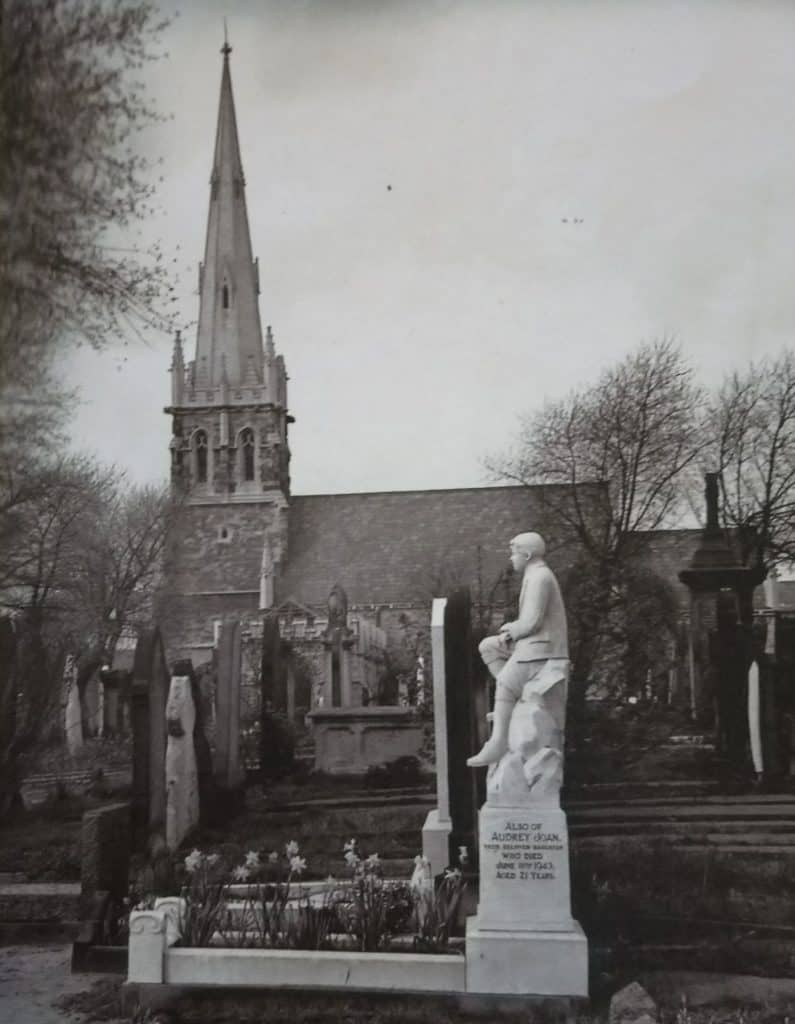 Black and white photo of Warstone Lane cemetery with white marble statue of boy in foreground and chapel in background.